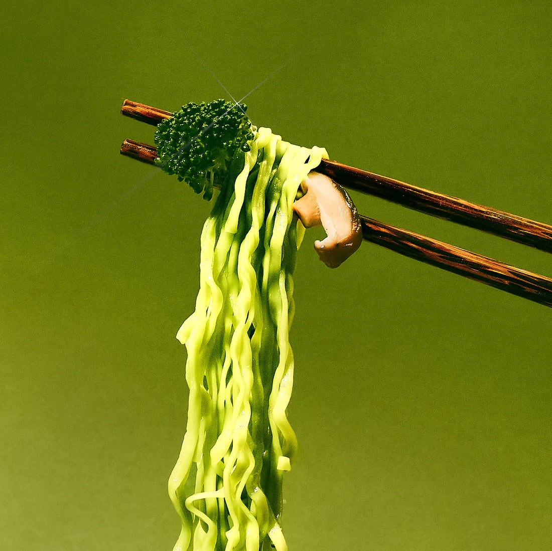 Chopsticks lifting green noodles with broccoli and mushrooms on a green background.