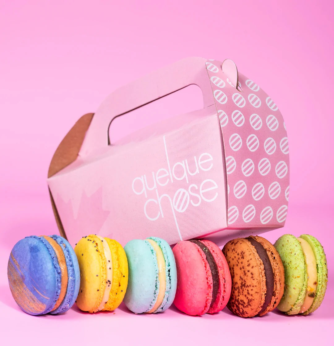 Row of colorful macarons beside a pink takeout box on a pink background.