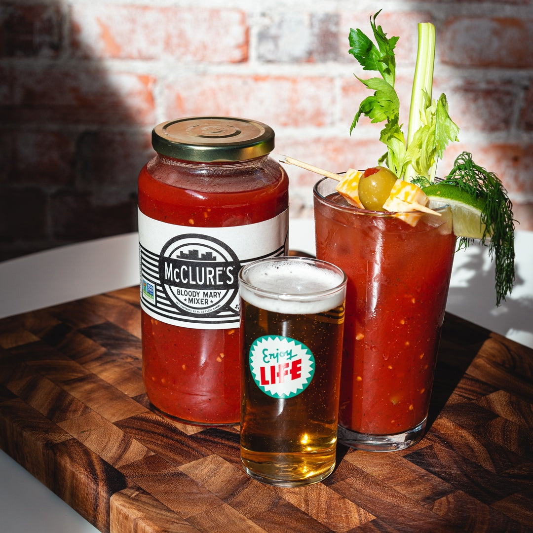 A jar of McClure's bloody mary mix, a mixed drink, and a beer are arranged on a wooden board.