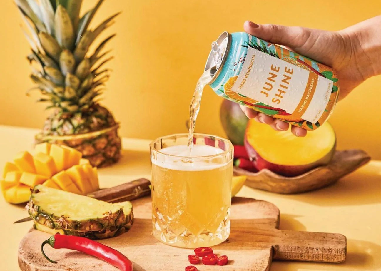 Hand pouring Juneshine Mango Daydream kombucha into a glass beside pineapple and mango.