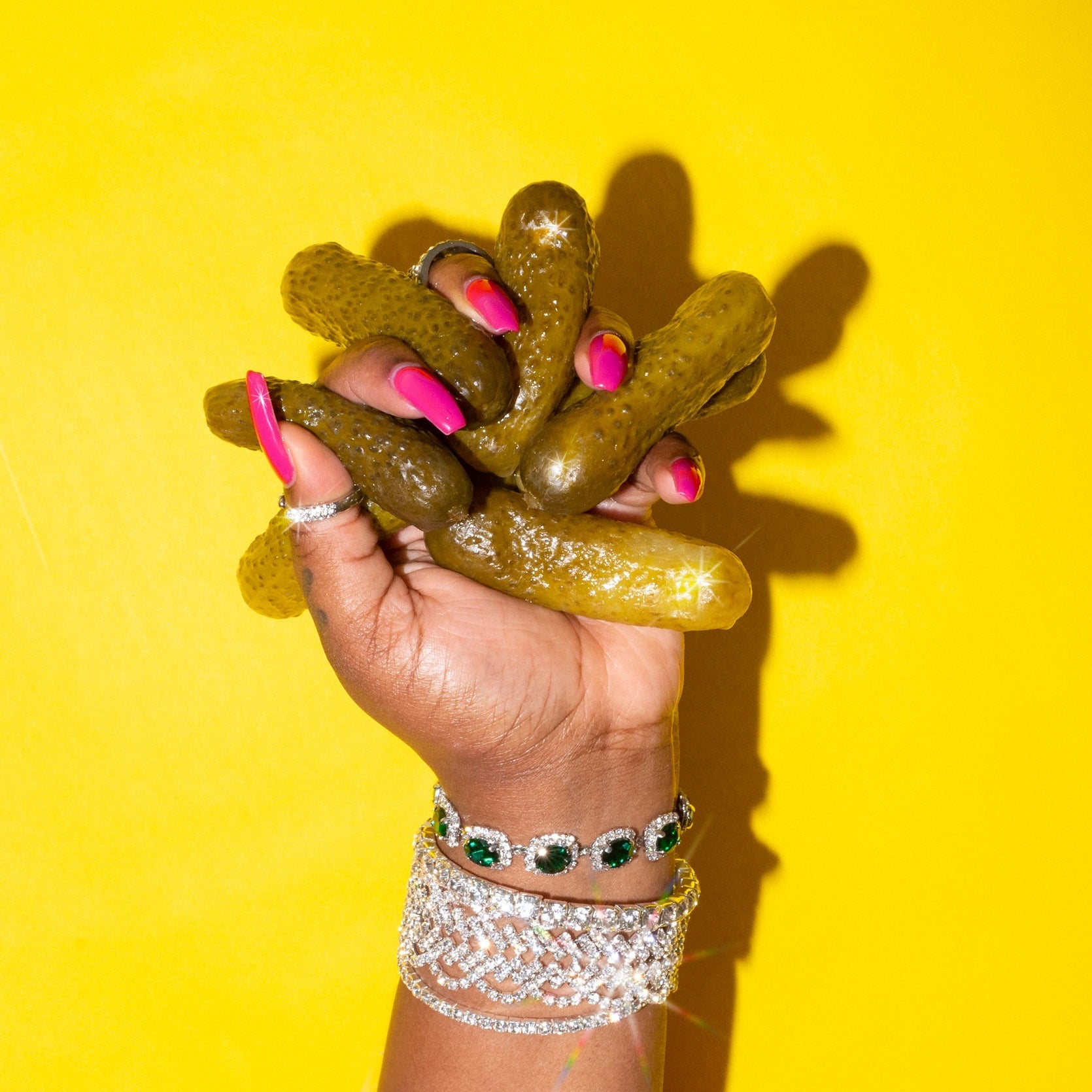 Hand with pink nails and silver jewelry holding pickles against a yellow background.