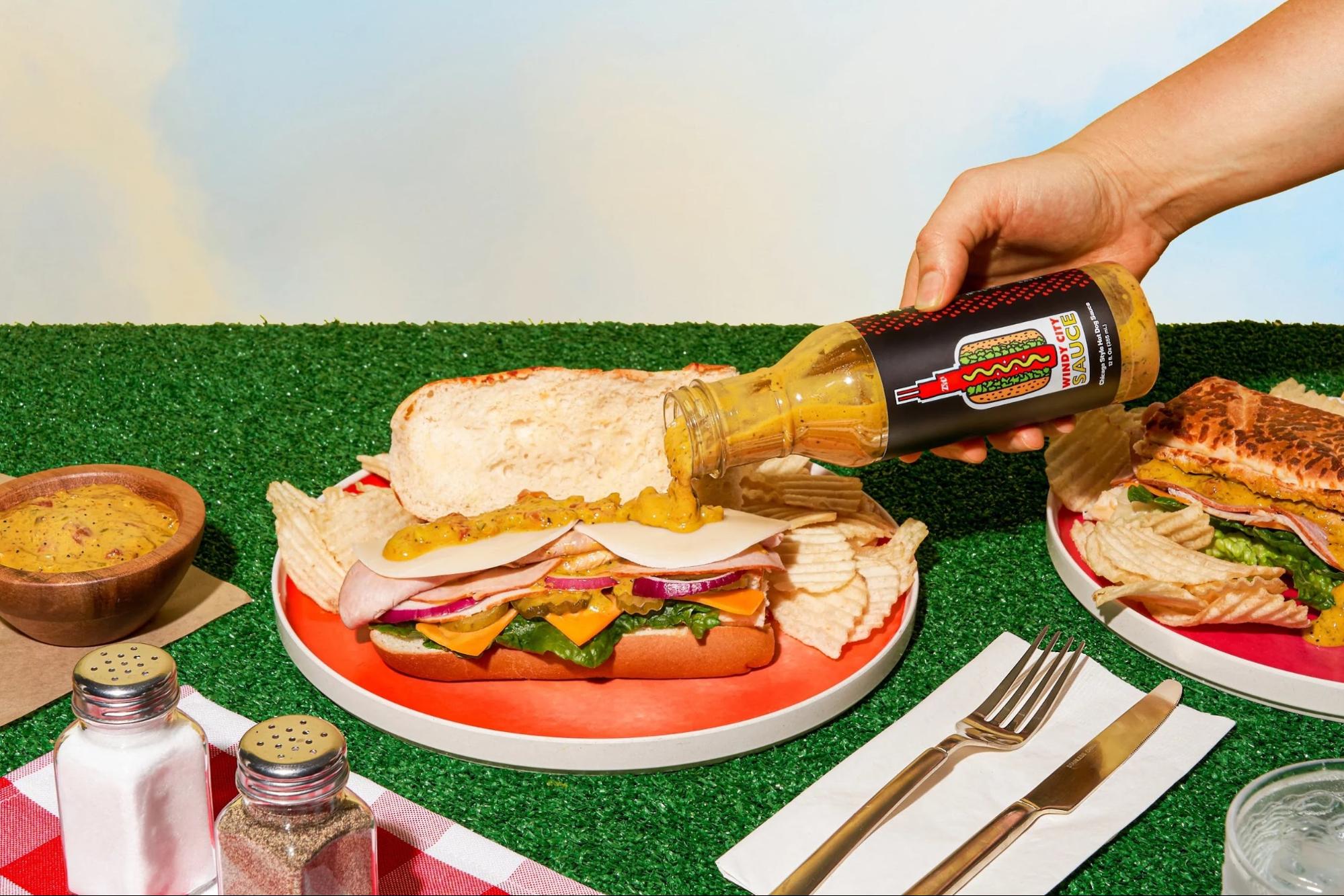 Hand pouring Windy City Sauce on a sandwich at a picnic setup with chips and mustard.