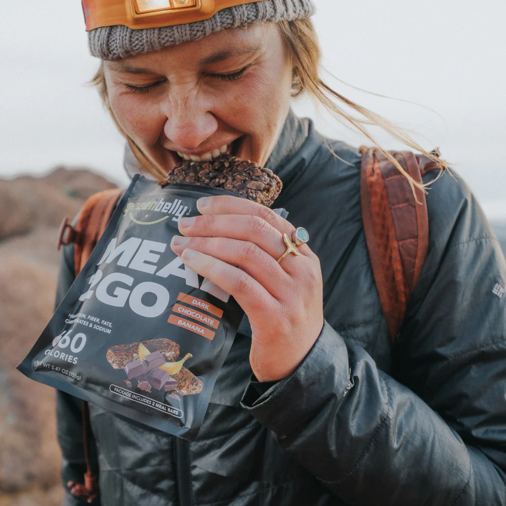 Hiker wearing outdoor gear eats a Greenbelly Meal2Go dark chocolate banana bar while on a trail.