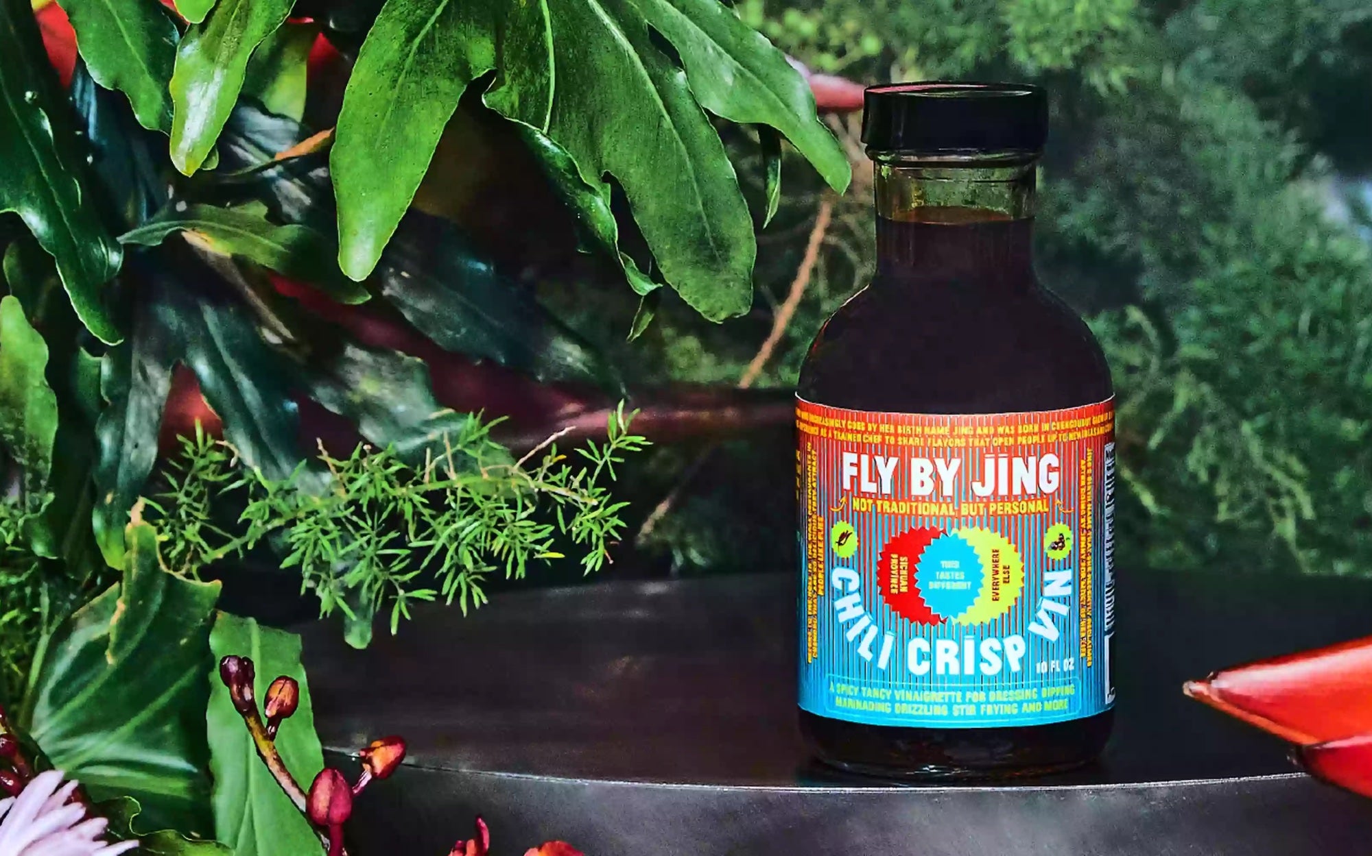 Bottle of Fly By Jing Chili Crisp Vinaigrette surrounded by green leaves and herbs.
